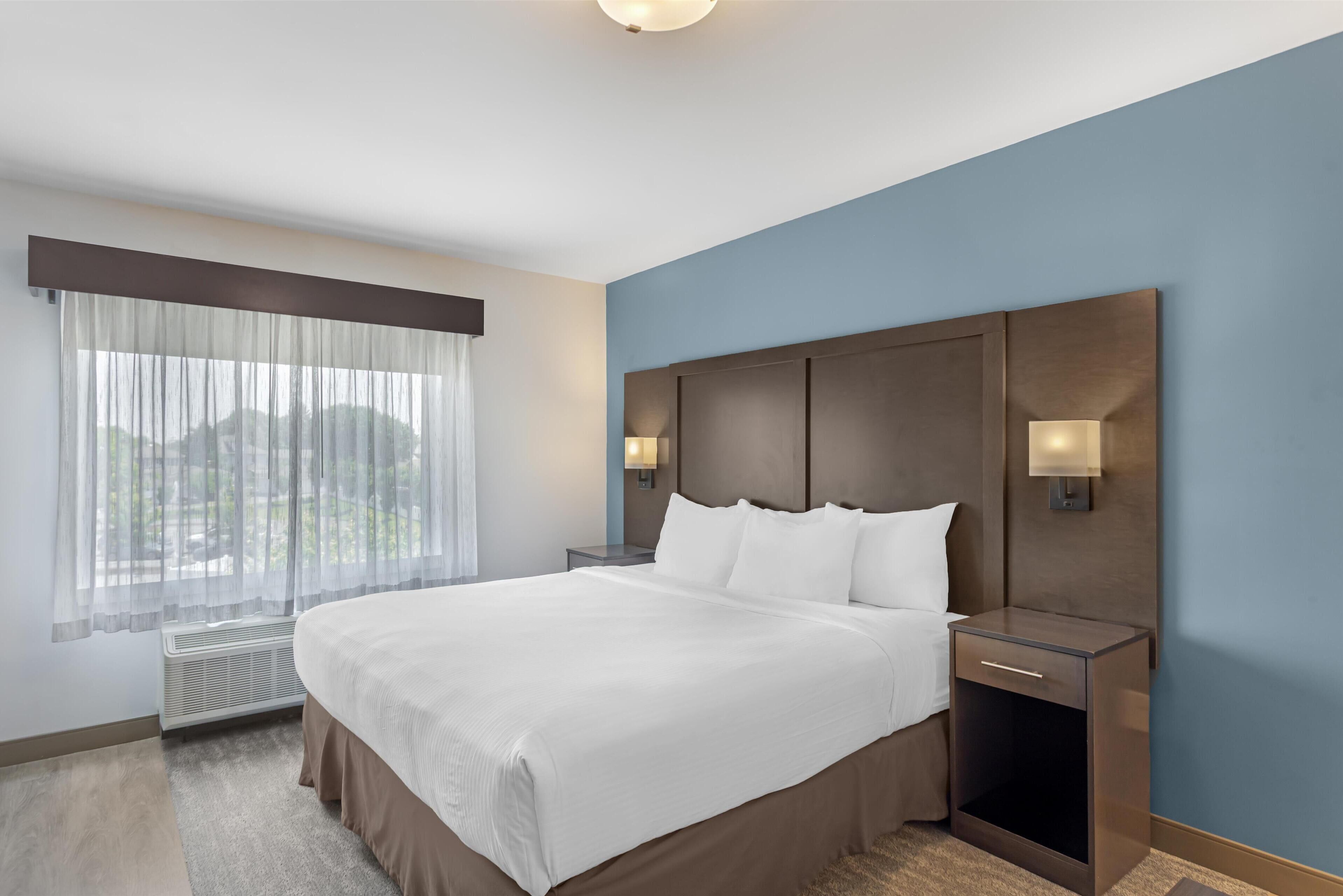 A hotel room with a large bed, a nightstand with a lamp, a window with sheer curtains, and a wall-mounted light fixture.