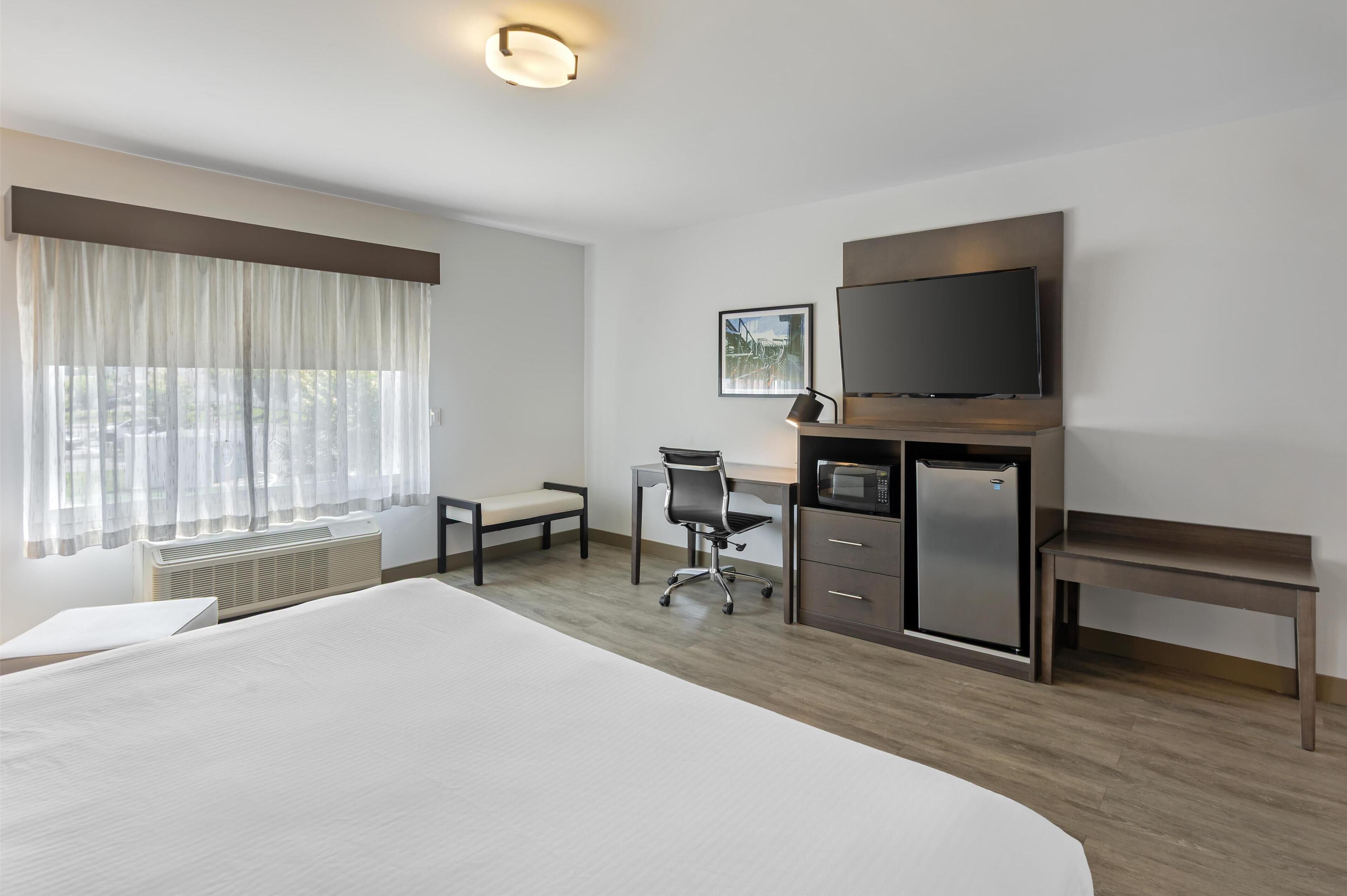 Quarto de hotel com cama grande, escrivaninha com cadeira, televisão, micro-ondas e um banco.