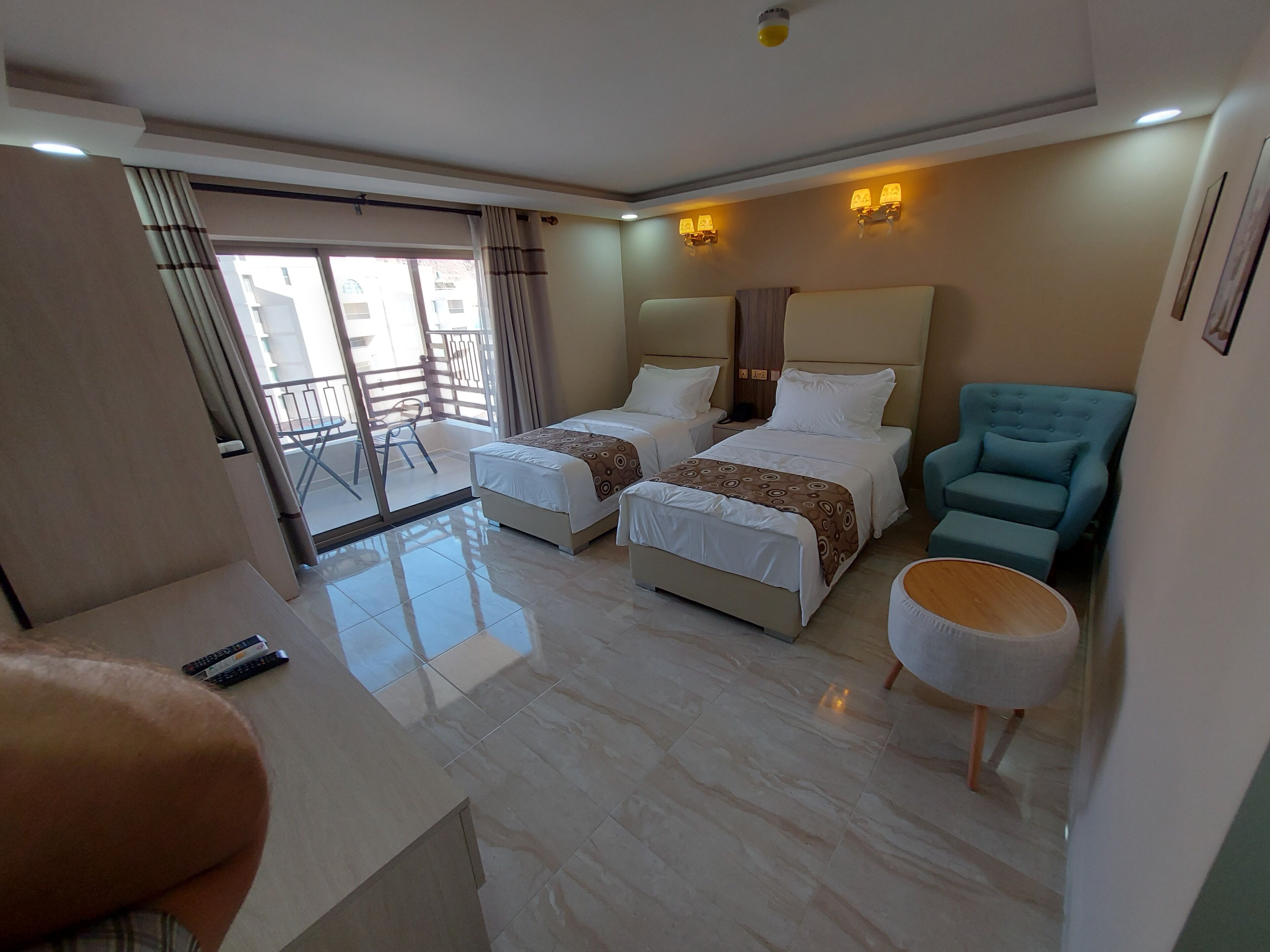 A hotel room with two beds, a blue armchair, a round wooden table, and a balcony with a view.