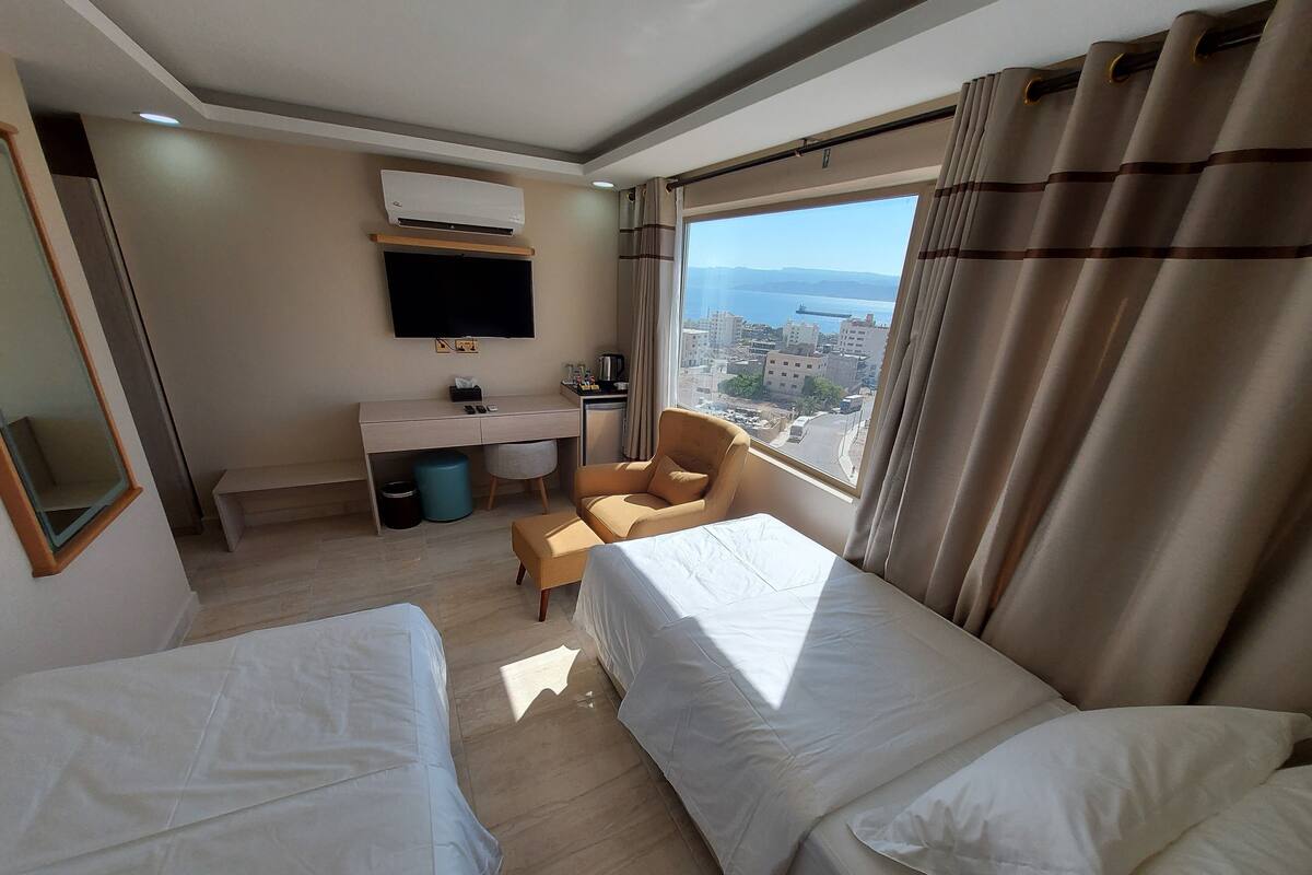 A hotel room with two beds, a desk, a chair, a TV, and a view of the city.