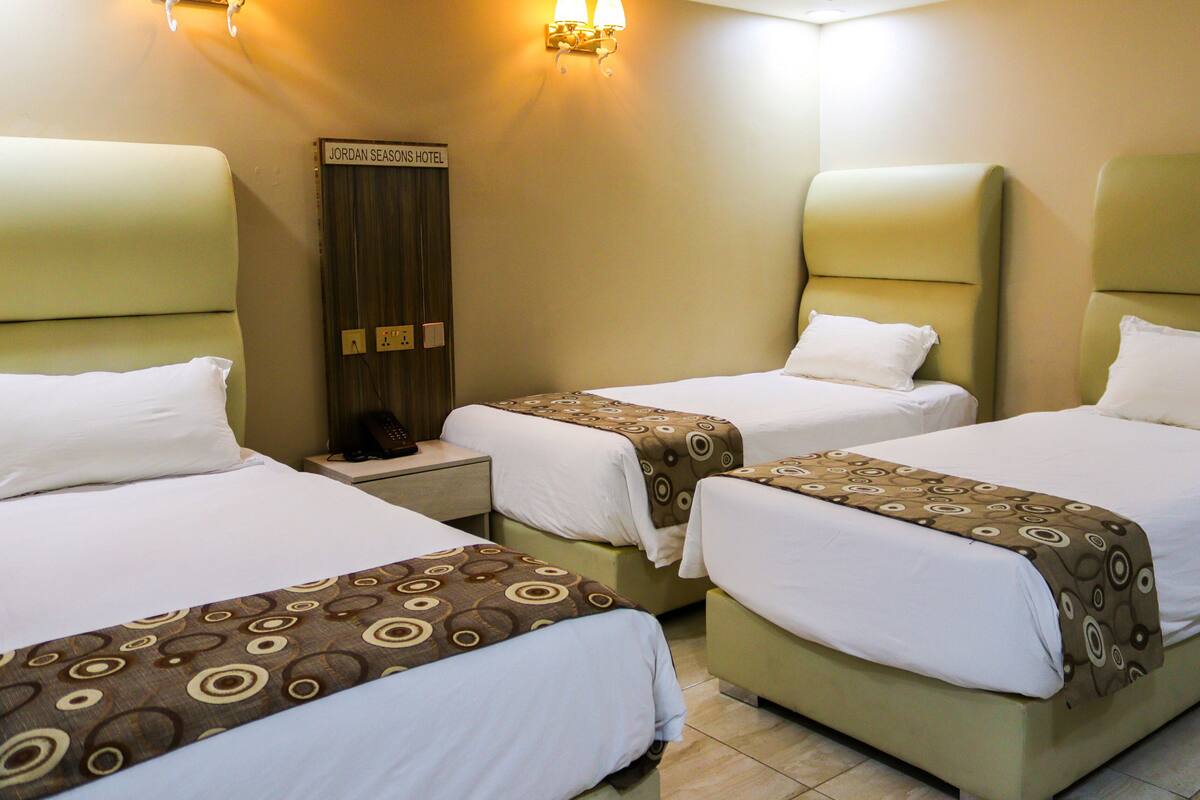 A hotel room with three beds, each with a headboard and a bedside table. There is a sign on the wall above the beds that reads "JORDAN SEASONS HOTEL".
