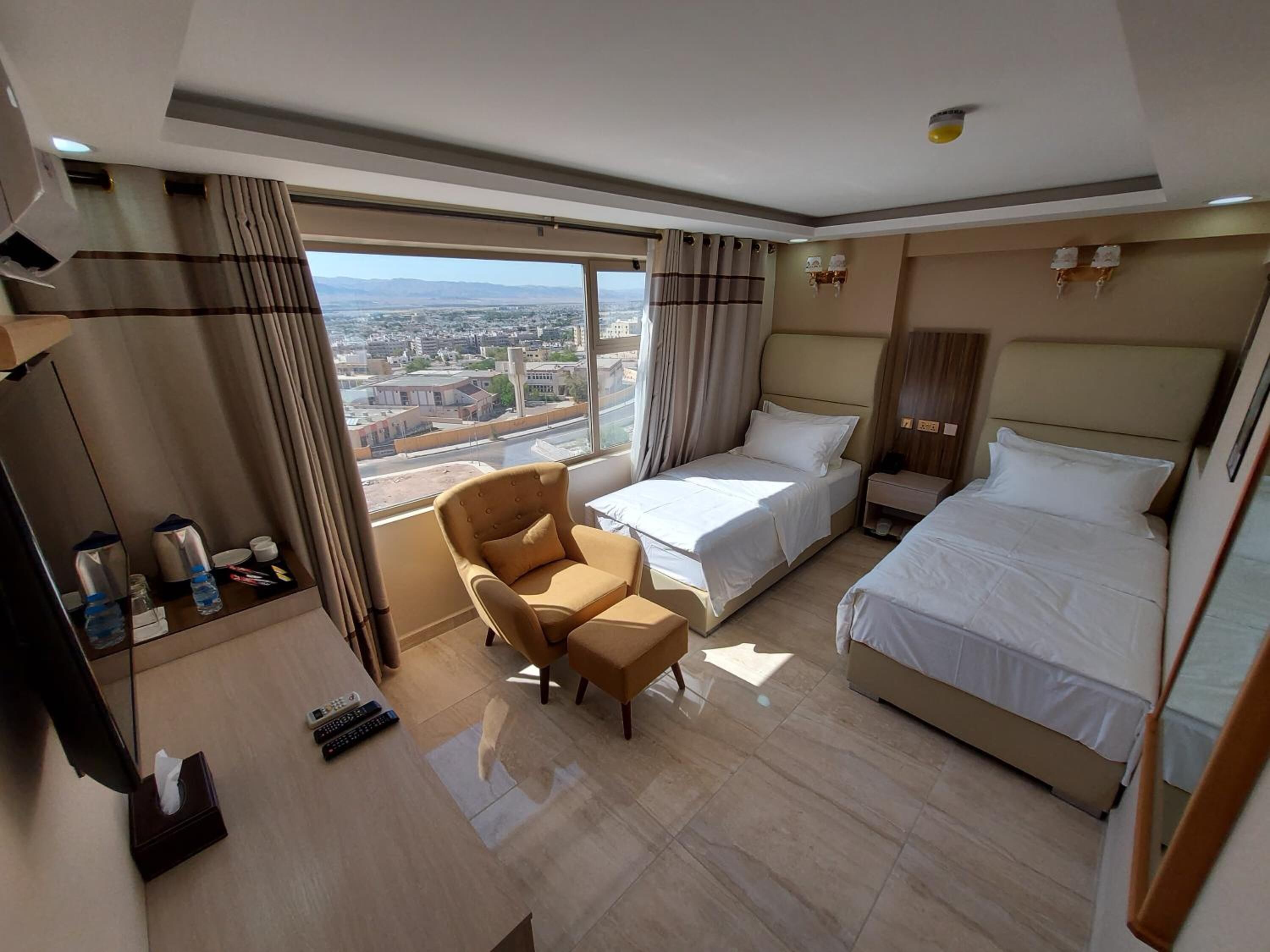 A hotel room with two beds, a sofa, a desk, and a large window offering a city view.
