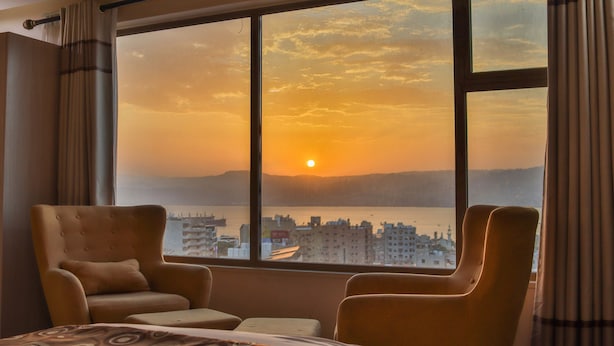 A hotel room with a large window offering a view of a cityscape and a sunset. - Jordan Seasons Hotel (Aqaba)