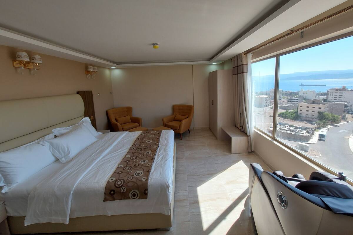 A hotel room with a bed, two armchairs, a wardrobe, and a large window with a city view.