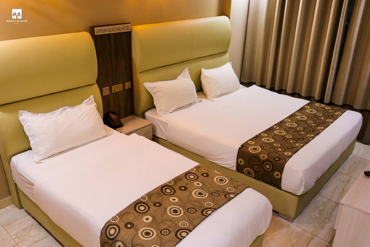 A hotel room with two beds, each with white linens and a brown patterned bedspread. There is a nightstand with a phone and a sign indicating the hotel name.