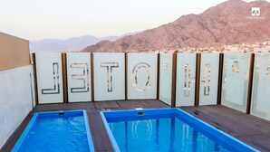 A rooftop pool area with two rectangular pools and a view of a cityscape and mountains in the background. - Jordan Seasons Hotel (Aqaba)