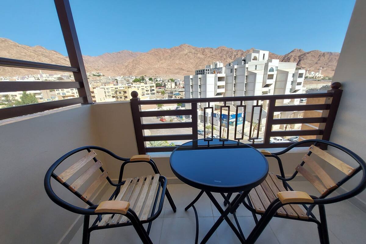 A balcony with a table and two chairs, overlooking a cityscape with mountains in the background.