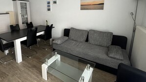 Un salon moderne avec un canapé gris, une table basse en verre, un coin repas avec une table et des chaises noires, et un meuble avec des portes en verre.