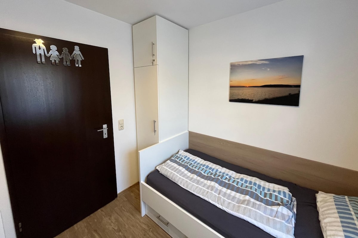 A bedroom with a bed, a wardrobe, a door with a light switch, and a wall-mounted picture.