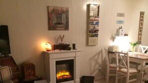 A cozy room with a fireplace, a dining table with chairs, a television, and a bed with a floral bedspread. - Ferienwohnung Hüttenzauber (Sankt Englmar)