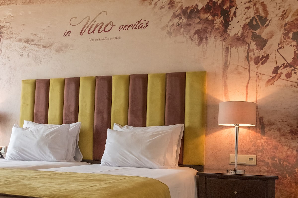 Une chambre d’hôtel comprenant un lit, des oreillers, une table de chevet avec une lampe, et un mur orné d’un motif texturé et d’un texte.