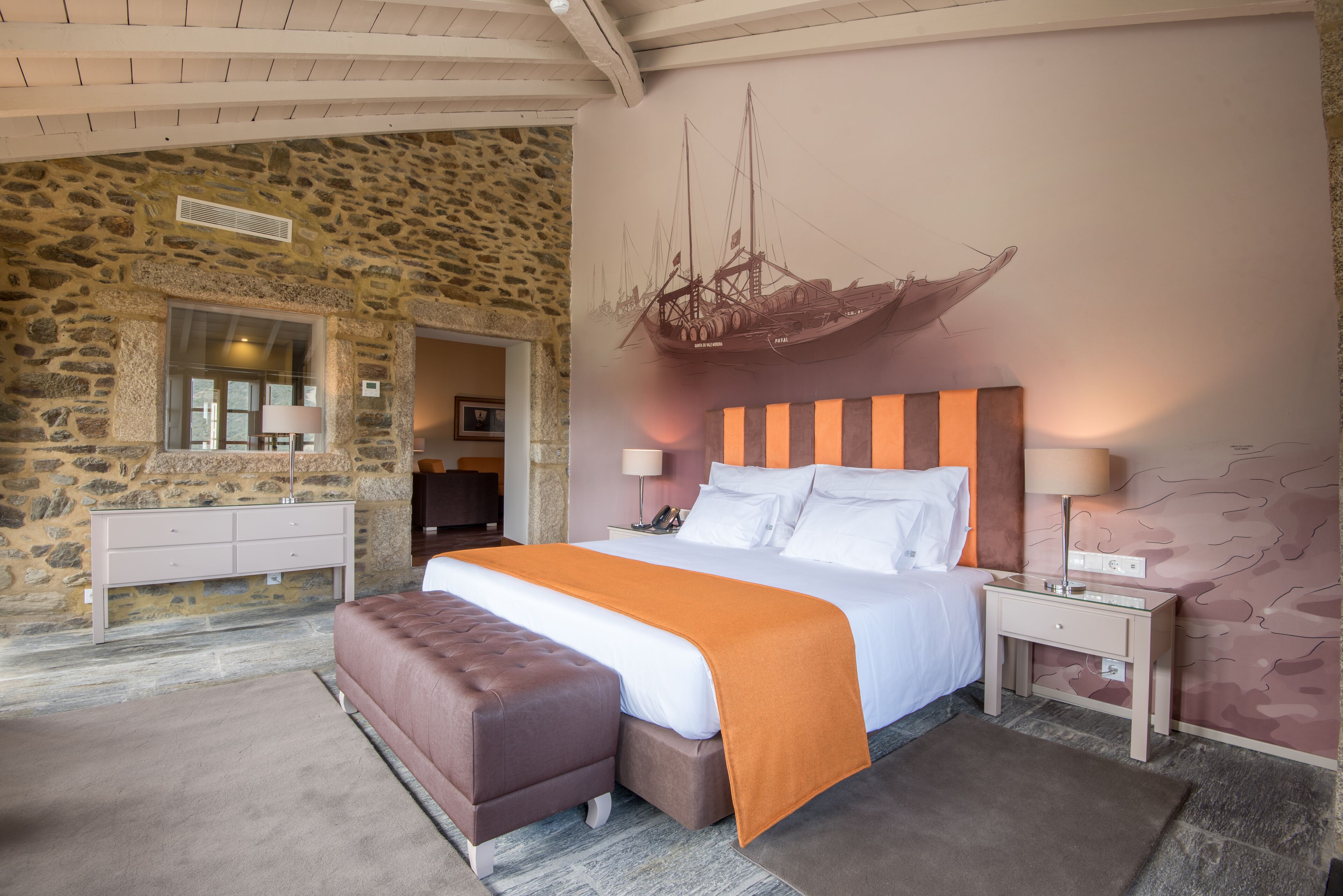 A bedroom with a stone wall, a bed with an orange blanket, a wooden headboard, and a nightstand with a lamp.