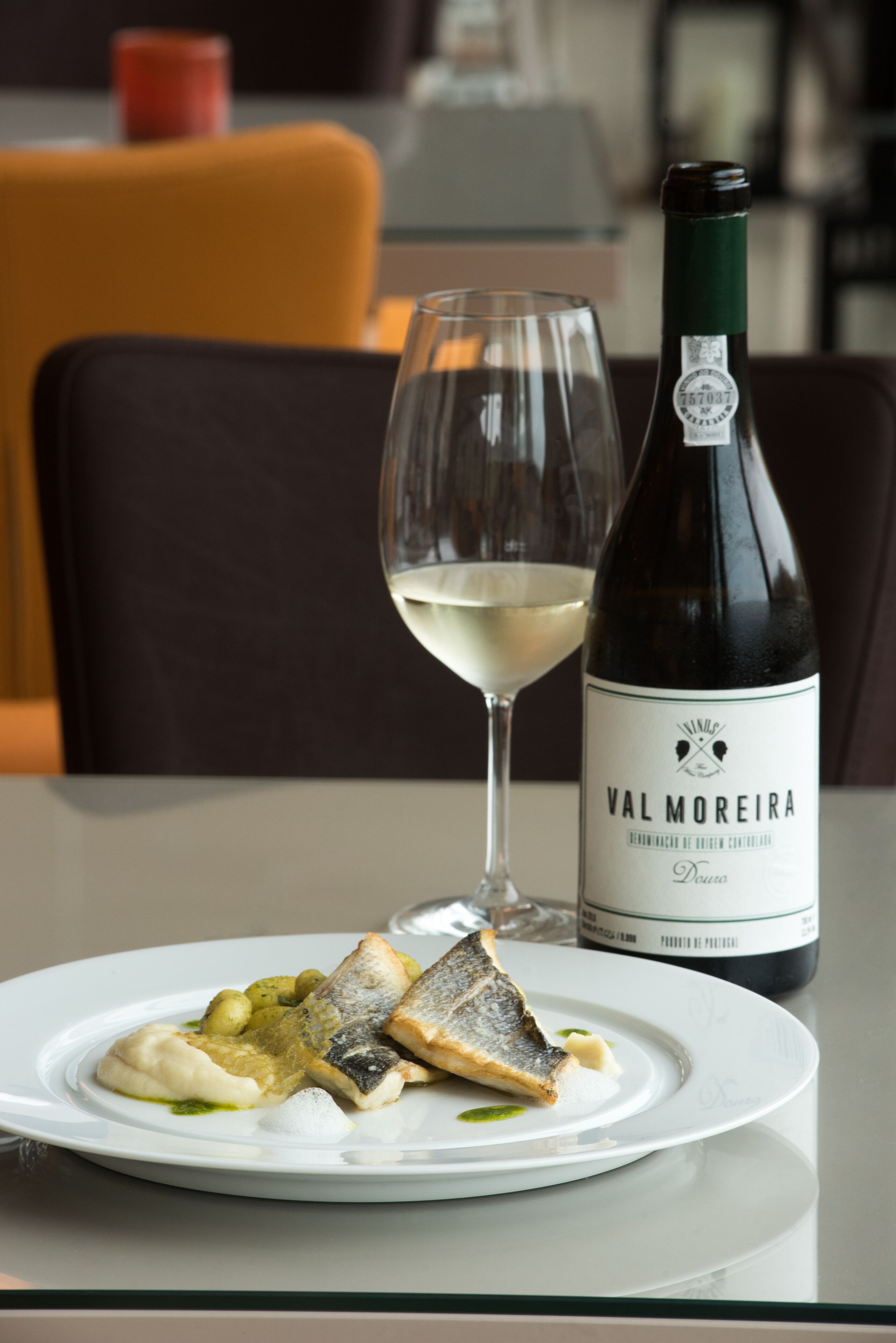 A plate of food with fish and potatoes, a glass of white wine, and a bottle of Val Moreira wine on a table.