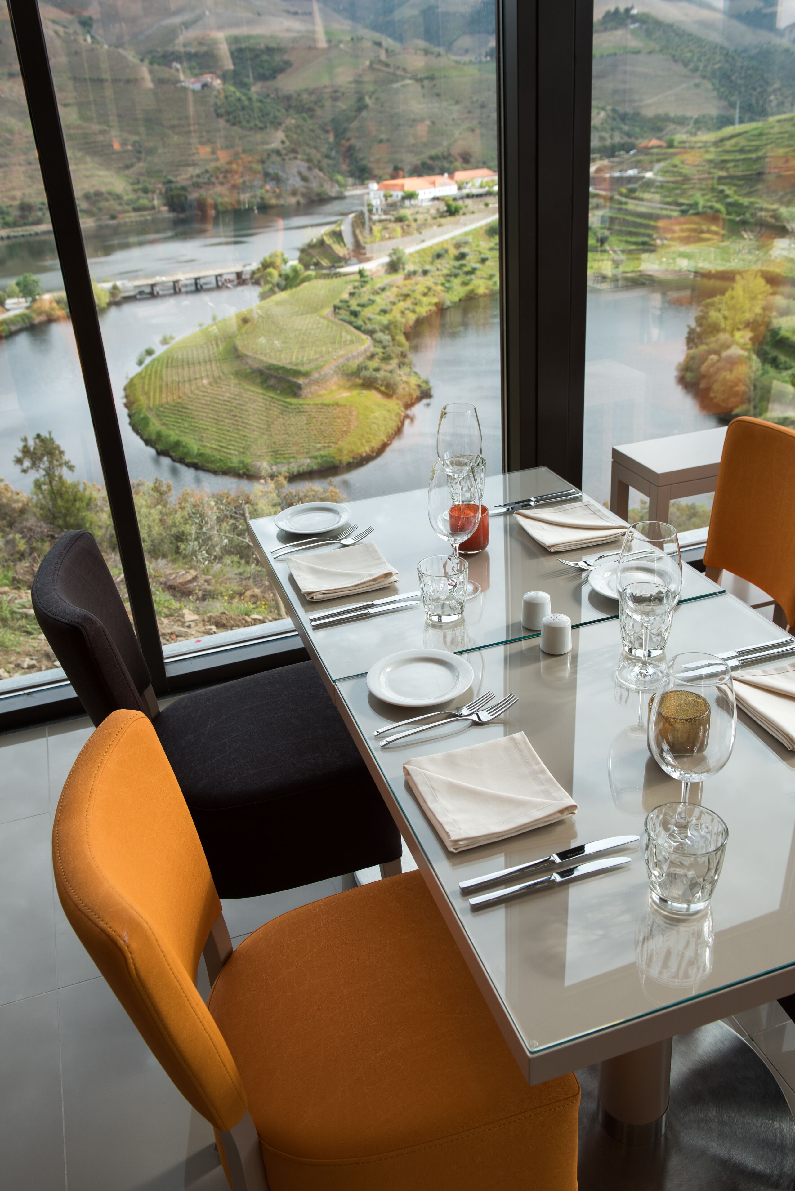 Una mesa de comedor dispuesta con platos, vasos y cubiertos, con vistas a un río y un paisaje montañoso.