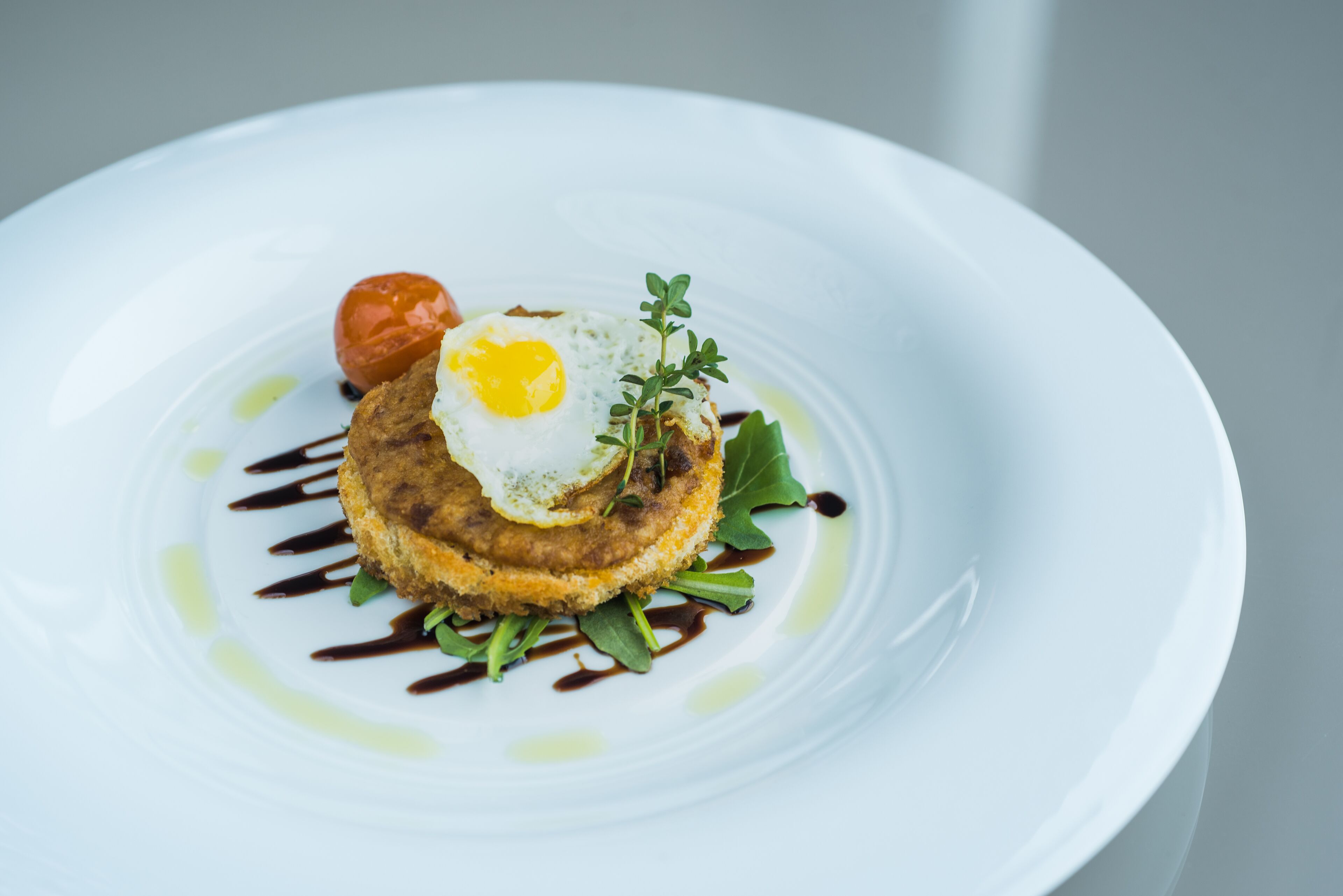Plato blanco con un plato de comida que incluye un alimento frito, un huevo frito y un tomate pequeño, decorado con hojas verdes y salsa.