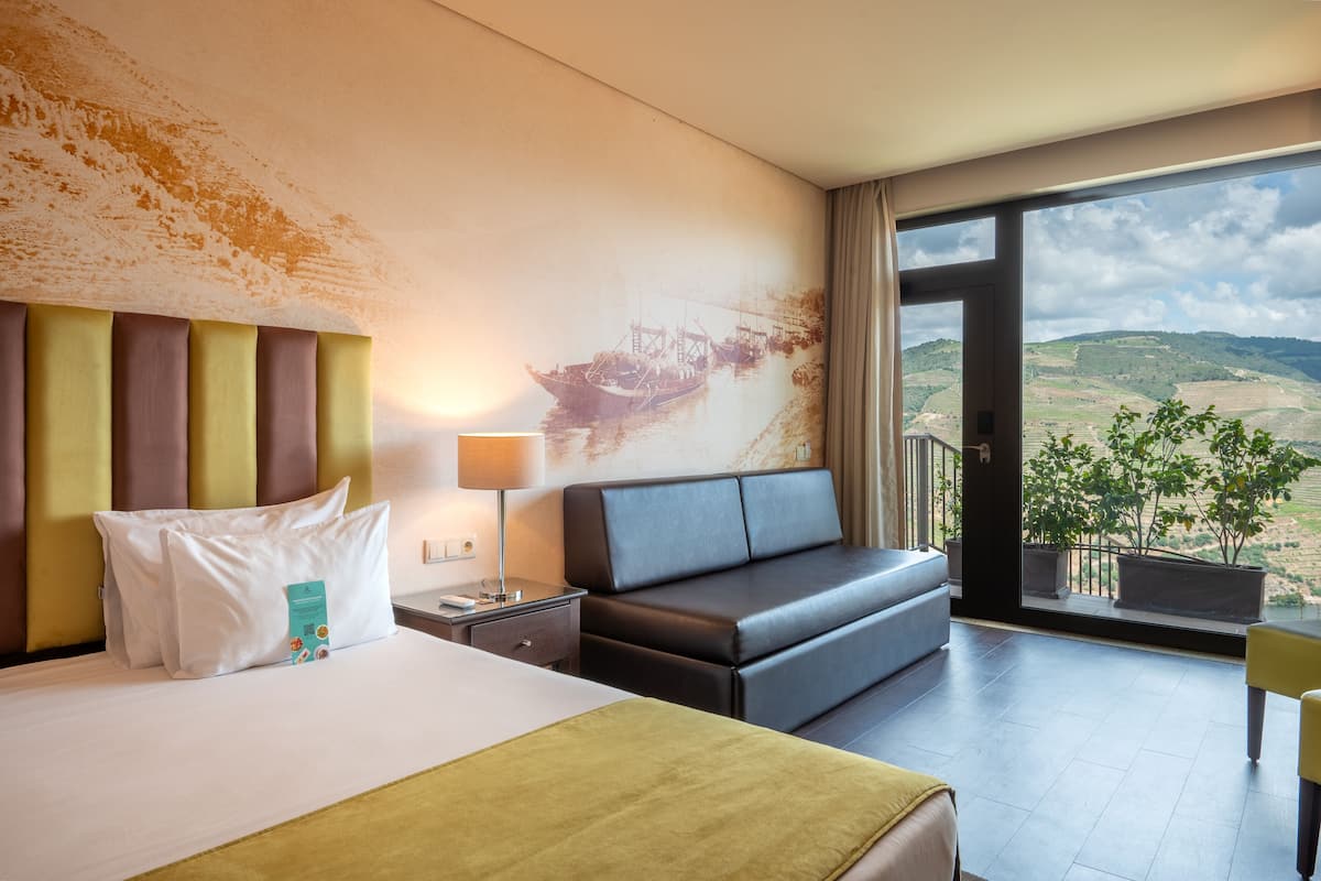 Une chambre d’hôtel avec un grand lit, un canapé, une table de chevet, une lampe et une vue sur la campagne, visible à travers une grande fenêtre.