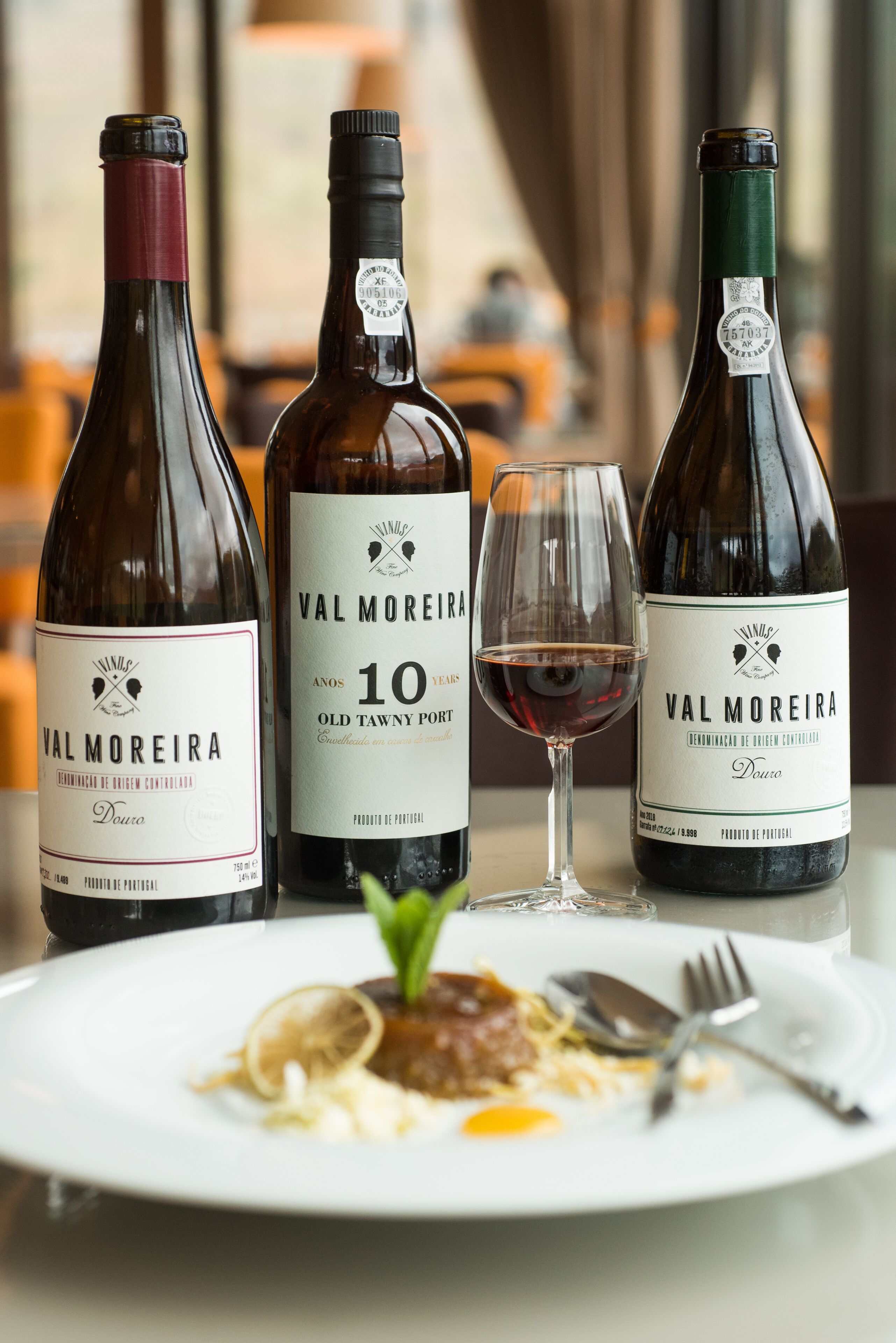 Three bottles of Val Moreira wine, a glass of red wine, and a plate of food on a table.