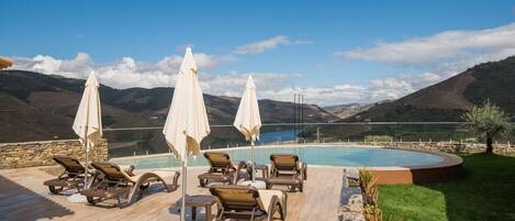 Zona de piscina con sillones de descanso y sombrillas, rodeada por un muro de piedra y vistas a un lago y montañas.