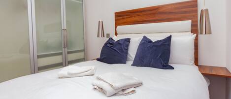 Una cama bien hecha con sábanas blancas y almohadas azules, un cabecero de madera y una mesita de noche con una lámpara.