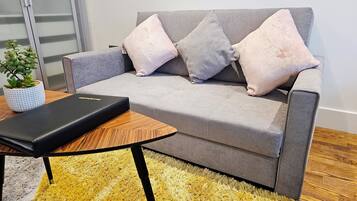 En grå sofa med tre puder, et lille træsidebord, en sort bog og en gul og grå lurvet måtte.