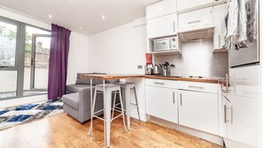 Een moderne keuken met witte kasten, een magnetron en een koelkast. Er is een eethoek met een tafel en twee barkrukken. Een grijze bank staat bij het raam, dat paarse gordijnen heeft.