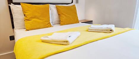 A neatly made bed with white and yellow bedding, a wooden headboard, and a bedside table with a lamp.