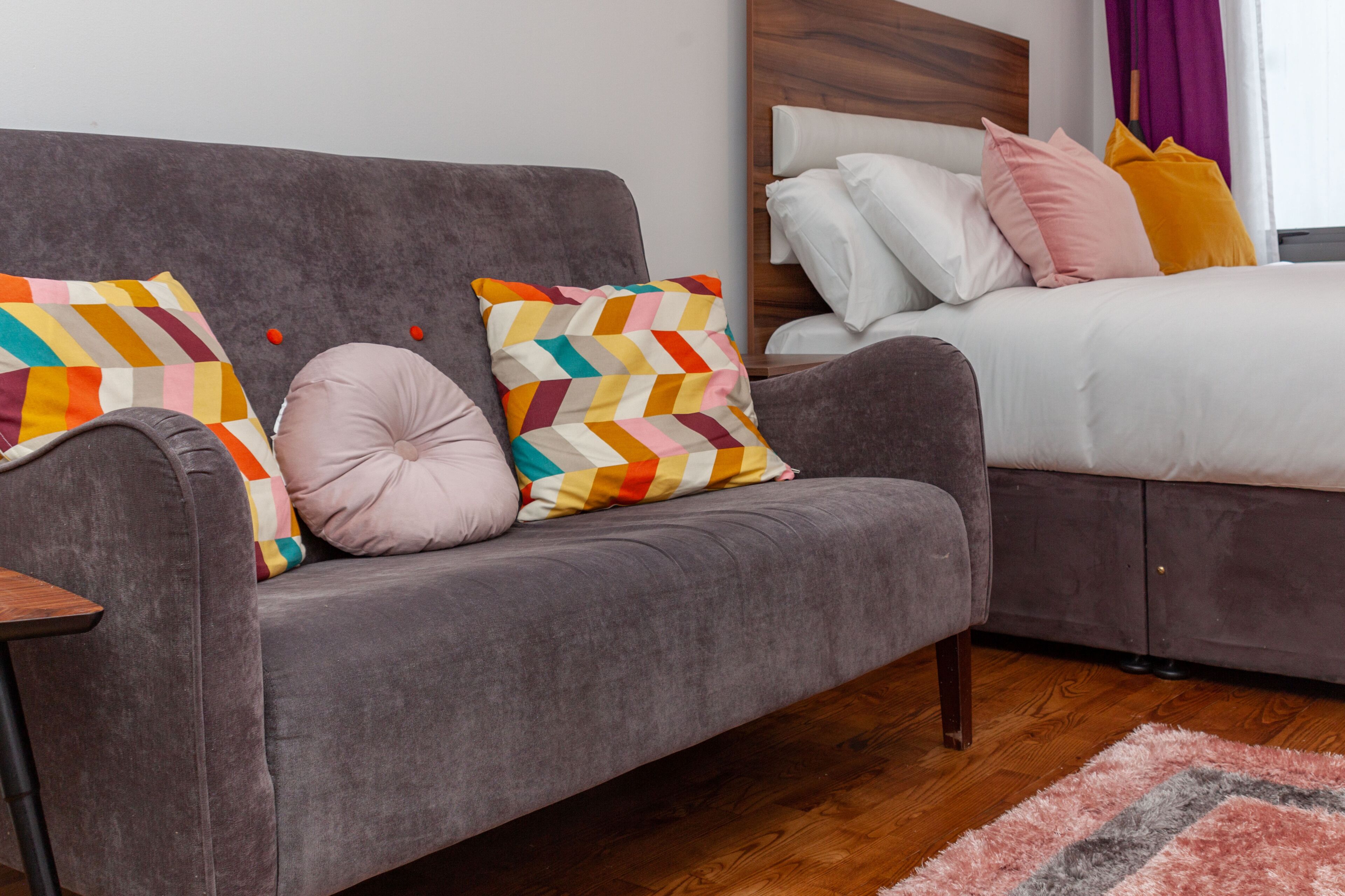 Un dormitorio moderno con un sofá gris, una cama con cabecera de madera y un cojín de estampado geométrico colorido.