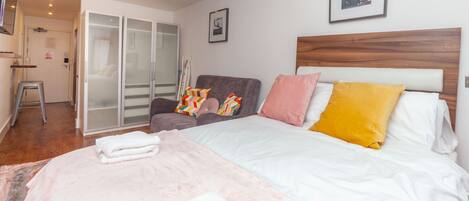 A bedroom with a bed, a grey armchair, a wooden headboard, and a white wall with a framed picture.