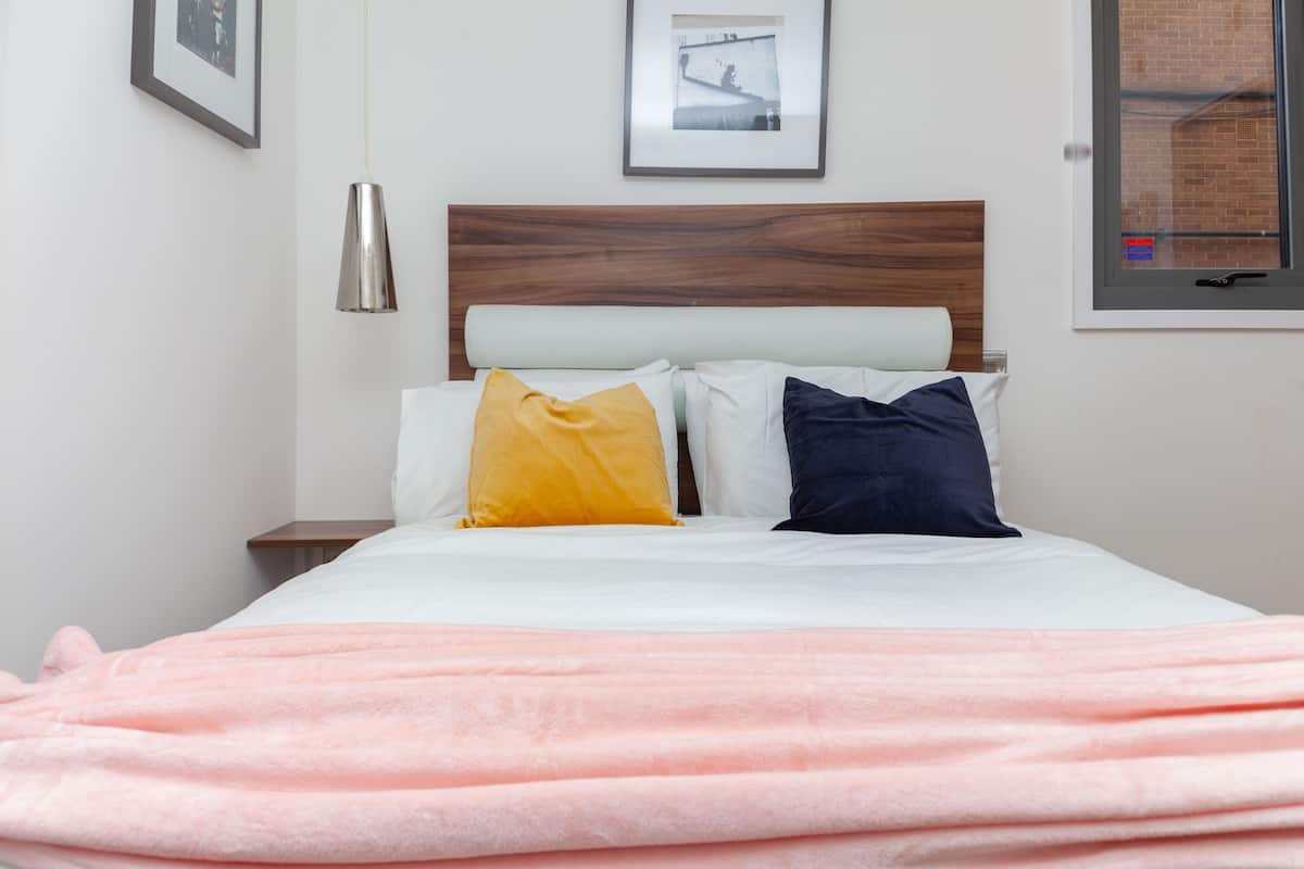 A neatly made bed with a pink blanket, a wooden headboard, and two pillows (one yellow, one blue).