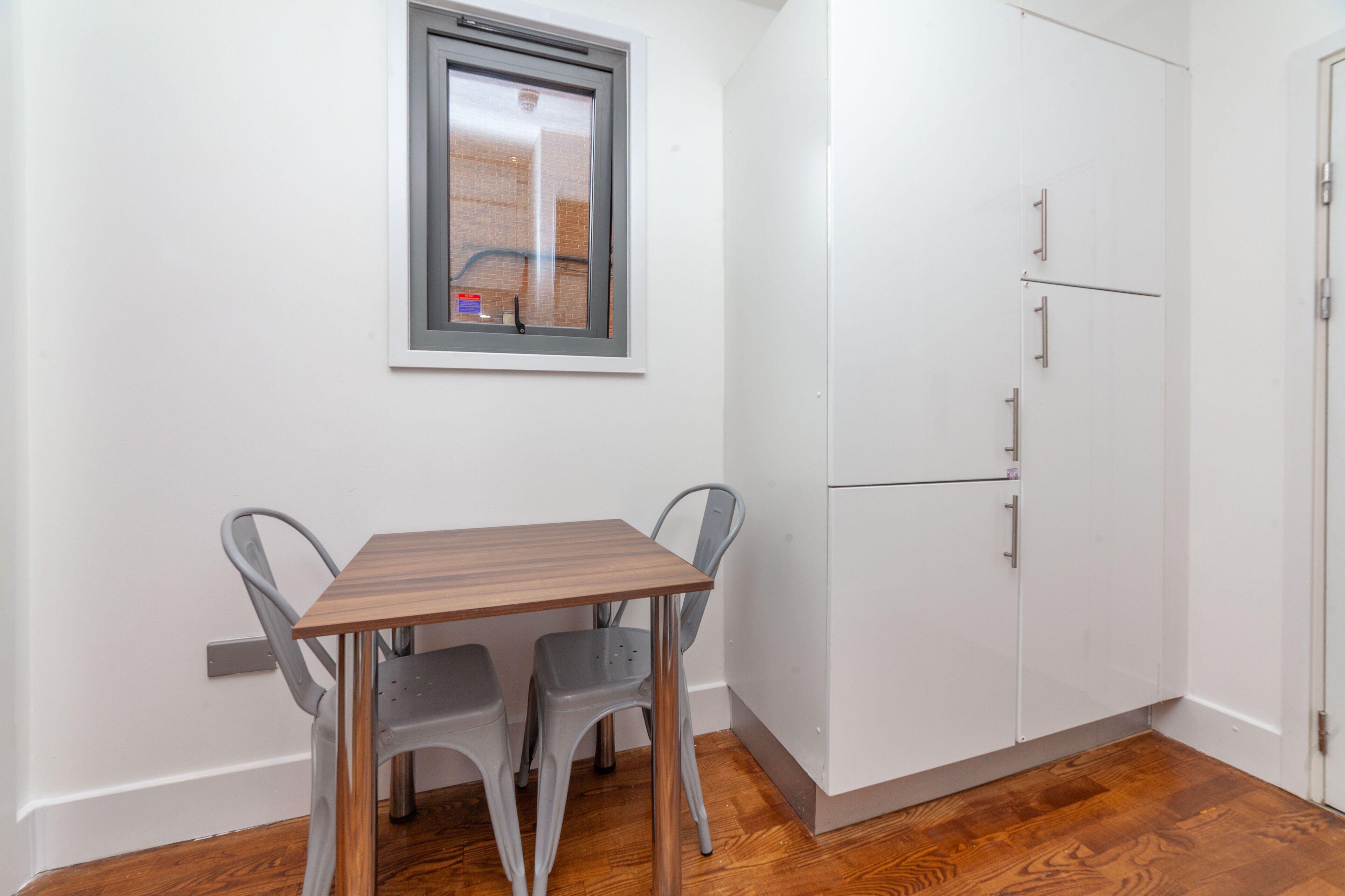 Una pequeña cocina con una mesa de madera y dos sillas, una ventana y armarios blancos.