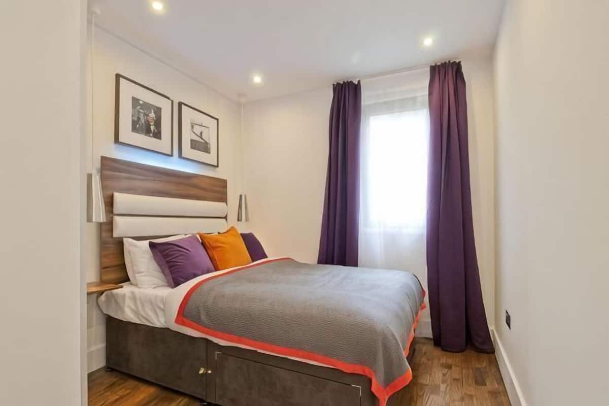 A bedroom with a bed, headboard, wall-mounted lights, and a window with curtains.