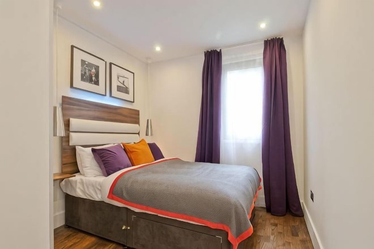 A bedroom with a bed, headboard, wall-mounted lights, and a window with curtains.