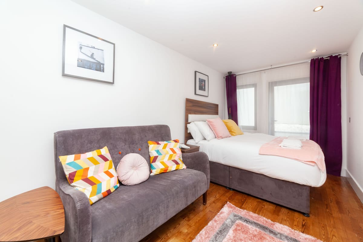 A modern hotel room with a grey sofa, a bed with a wooden headboard, a small wooden side table, and a large window with purple curtains.