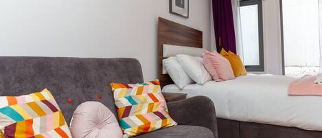 A modern hotel room with a grey sofa, a bed with white bedding, colorful pillows, and a wooden headboard.