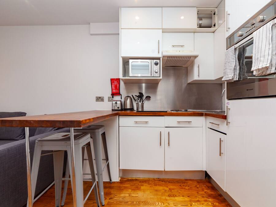 Cozinha compacta com armários brancos, micro-ondas e fogão. Há dois banquetes e uma mesa de madeira.