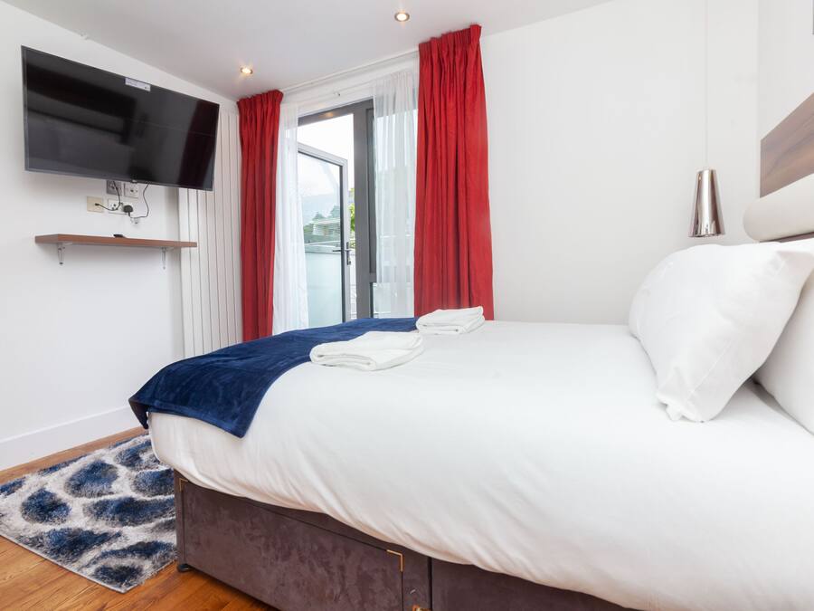 A modern hotel room with a large bed, a flat-screen TV mounted on the wall, and a sliding glass door with red curtains.