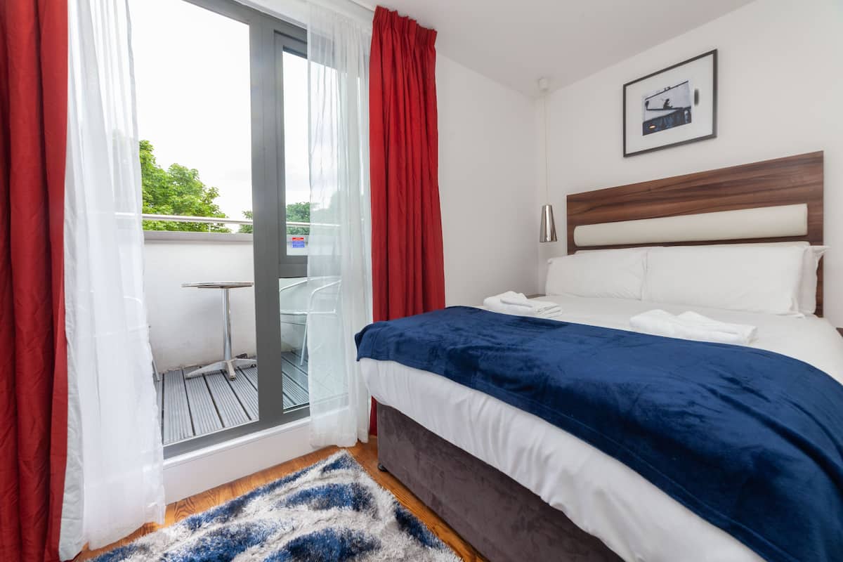 A bedroom with a bed, a window with red curtains, a white headboard, and a framed picture on the wall.
