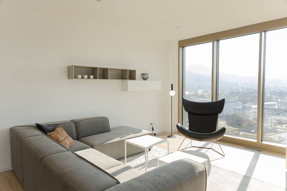 Uma sala de estar moderna com um sofá cinzento, uma poltrona preta, uma mesa de centro branca e uma grande janela com vista para a cidade.