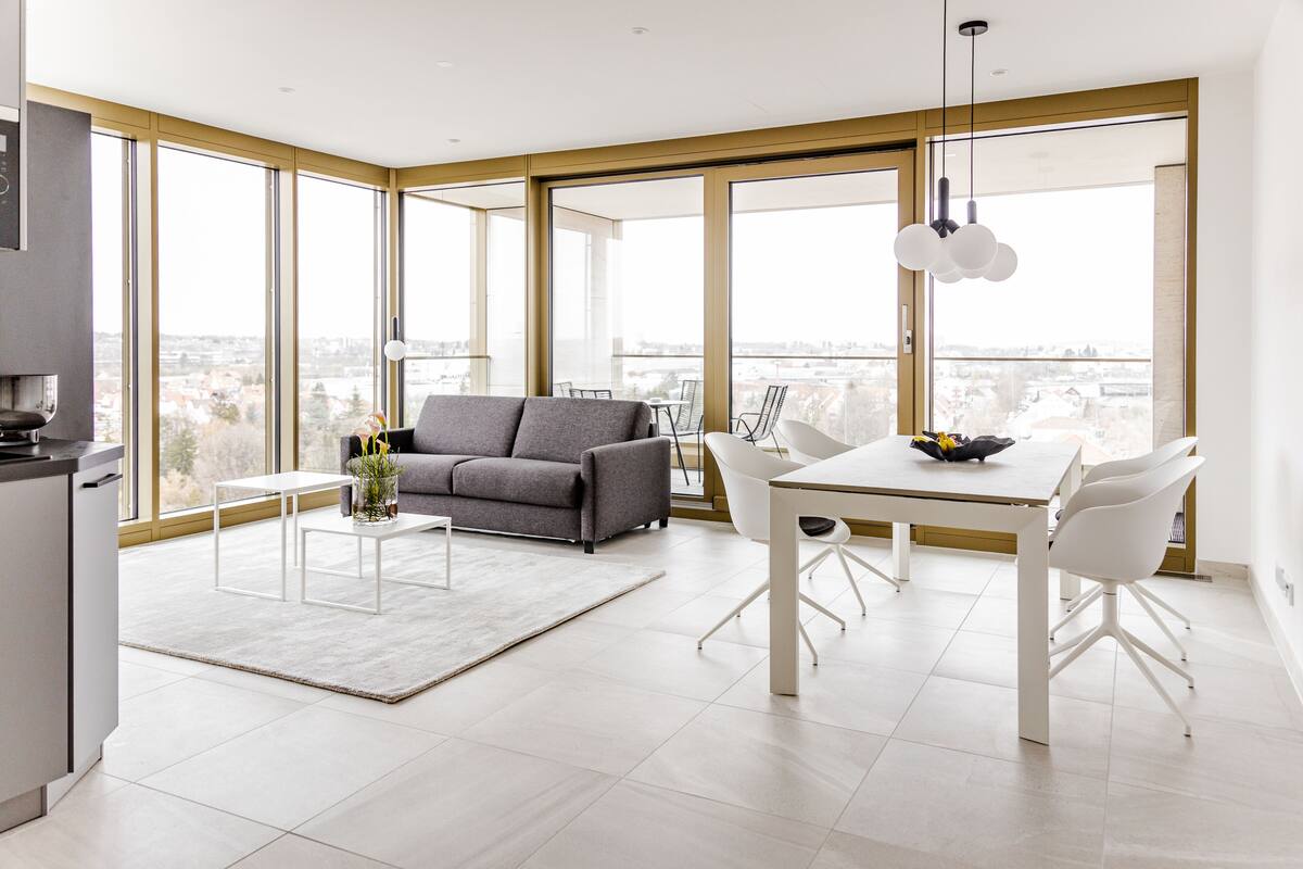 Uma sala de estar moderna com um sofá cinzento, uma mesa de jantar com cadeiras brancas e grandes janelas com vista para a cidade.