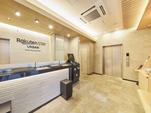 Una recepción con un cartel que dice 'Rakuten Stay URBAN Shijo Kawaramachi'. Hay dos ascensores, un bote de basura y una pequeña zona de estar.