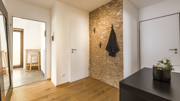 A modern room with a wooden floor, a brick accent wall, a black coat hanging, and a potted plant on a dark surface. - Ferienwohnung Whiskey (Winterberg)