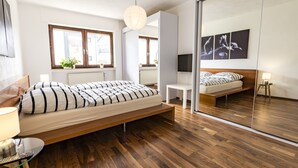 A modern bedroom with a large bed, bedside tables, a mirror, and a view of the city through large windows. - Ferienwohnung Whiskey (Winterberg)