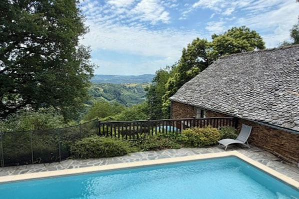 A swimming pool with a stone wall and a wooden deck, surrounded by greenery and trees.