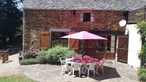 A stone building with a tiled roof, a covered patio with a table and chairs, and a garden area with flowers. - Converted Stone Barn with Pool (Occitanie)