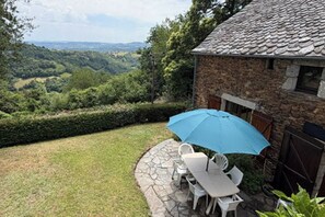 Un edificio de piedra con techo de tejas, un patio con sillas y mesa blancos, y una sombrilla azul.