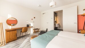 Et hotelværelse med seng, skrivebord, stol og et lille bord med en tilhørende stol. Der er et klædeskab, et spejl og en dør til et badeværelse.