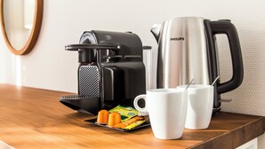 Kaffemaskine, elkedel og to hvide kopper på et træbord med kaffekapsler og en ske.