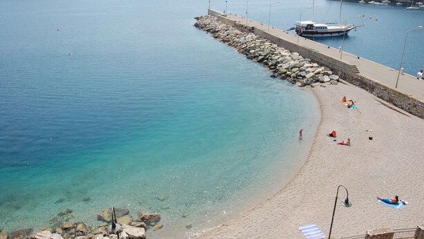 Una playa costera con un muelle de piedra que se adentra en el agua, mar turquesa transparente y algunas personas relajándose en la arena.