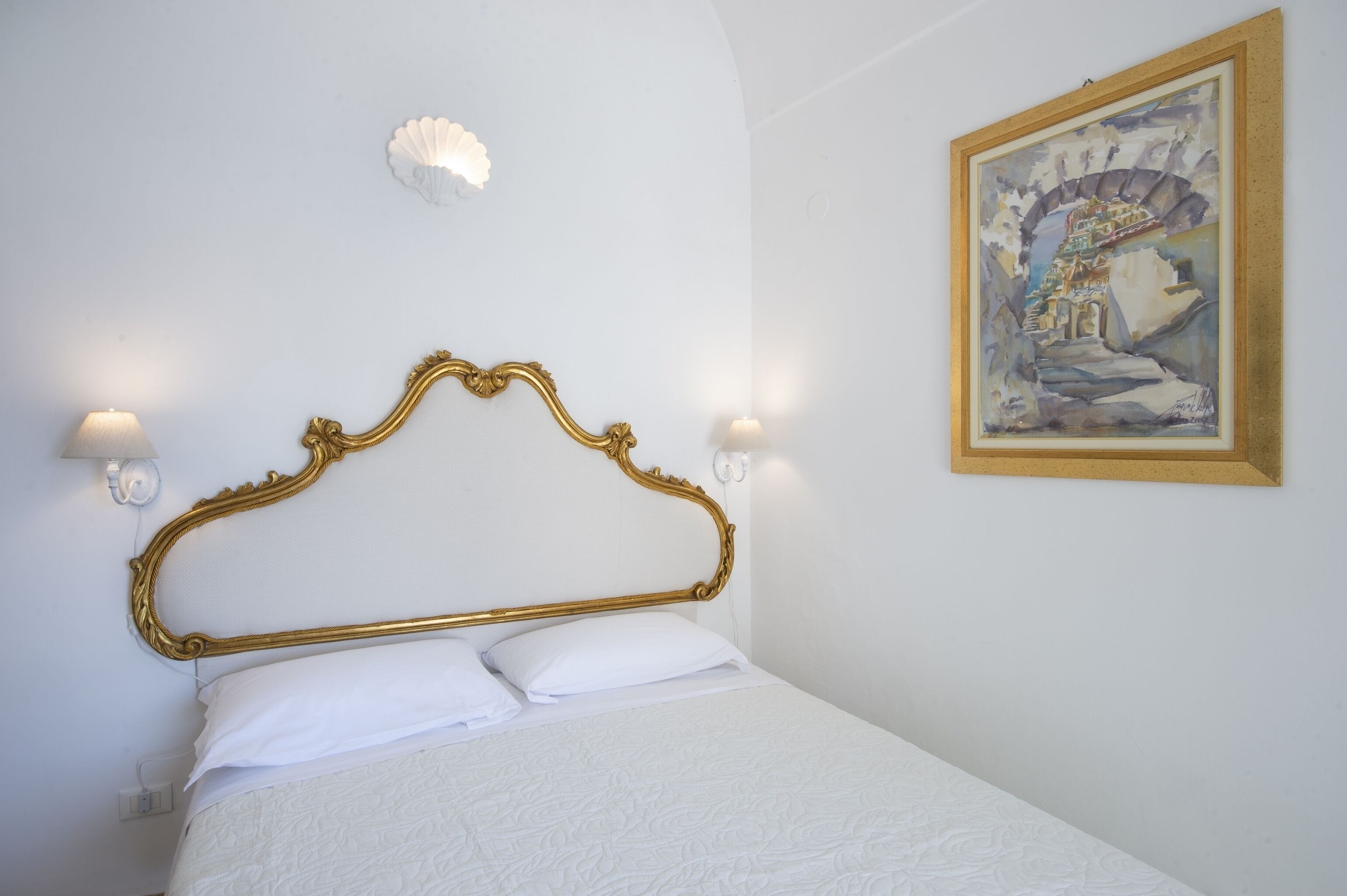 A neatly made bed with a gold headboard, two wall-mounted lamps, and a framed painting on the wall.