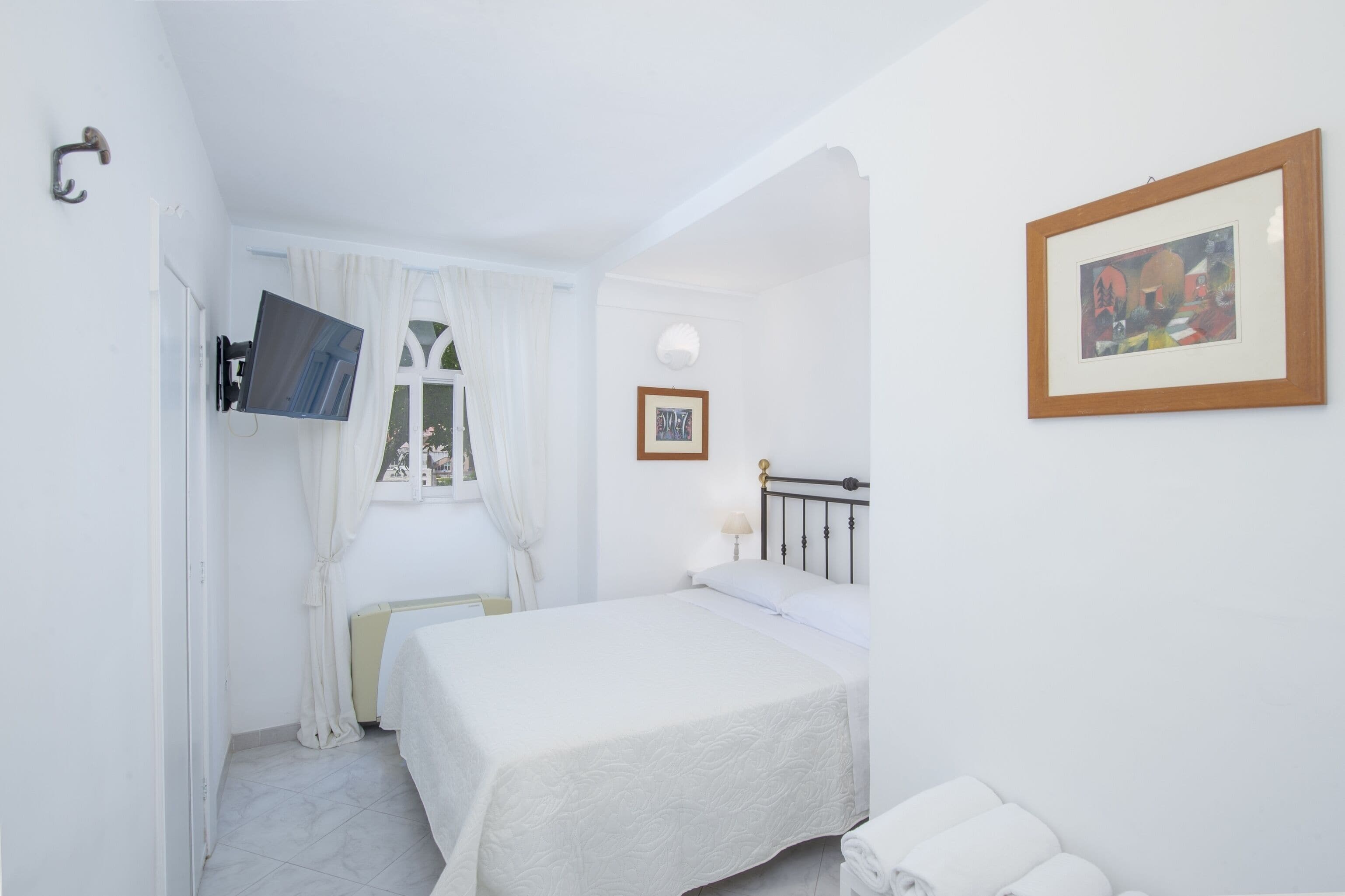 A bedroom with a bed, a TV mounted on the wall, a window with curtains, a framed picture on the wall, and a radiator.